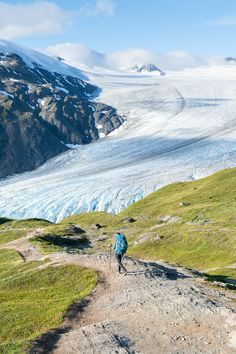 a person walking up a trail in front of a large ice cave and snow covered mountains