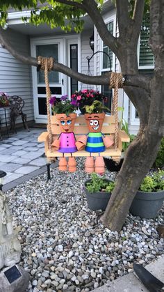 two wooden dolls are sitting on a swing in front of a tree with potted plants