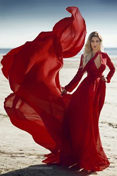 a woman in a red dress standing on the beach with her arms behind her back