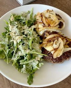 a white plate topped with meat covered in onions next to a green leafy salad