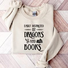 Easily Distracted By Dragons And Books Sweatshirt | Fantasy Book Lover Shirt | Reading Sweater | Book Crewneck | Dragon Book Sweatshirt Thank you for considering purchasing from Raven Printing Company.  We are based out of Cleveland, Ohio and would love to have the opportunity to earn your business. Because we are a small business, we cannot accept returns or exchanges.  However, if your product does not meet your standards please reach out and we would be more than happy to work with you on a s Literary White Cotton Tops, White Cotton Literary Tops, Bookish Cotton Sweatshirt With Letter Print, White Bookish Sweatshirt With Letter Print, Cotton Letter Print Bookish Sweatshirt, Literary Long Sleeve Cotton Tops, Sweatshirt And Shirt Outfit, Book Crewneck, Dragon Book