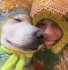 a white dog wearing a yellow hat and scarf next to a woman in a knitted sweater