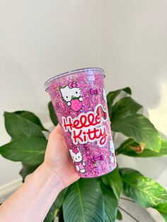 someone holding up a hello kitty cup in front of a potted plant with hello kitty on it