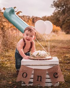 a young boy sitting on top of a table with balloons and a cake in front of him