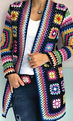 a woman wearing a colorful crochet jacket and jeans standing in front of a wall