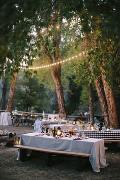 an outdoor dinner table set up with lights strung from the trees and picnic tables in the background