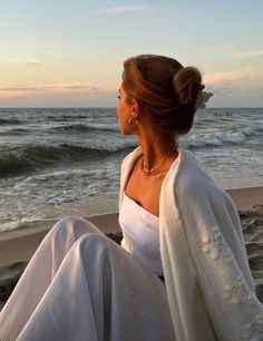 a woman sitting on top of a sandy beach next to the ocean in white clothes