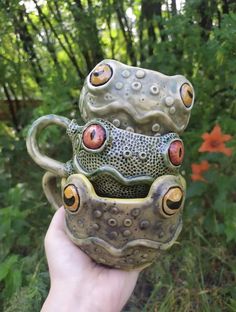 a hand holding two ceramic mugs in front of some bushes and trees with orange eyes