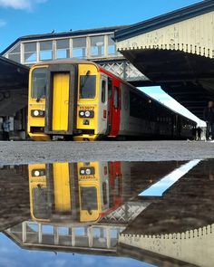 a yellow and red train is parked in front of a building with its reflection on the ground