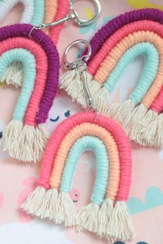 three rainbow shaped key chains with tassels hanging from them on a pink background