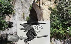 a sign with a witch on it in front of some trees and bushes, next to a rock wall