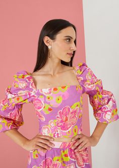 a woman wearing a pink floral dress with puffy sleeves and an open shoulder, standing in front of a pink wall