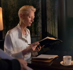 a woman sitting at a table with a book in her hands and a cup next to her