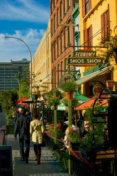 two people walking down the sidewalk in front of shops and restaurants on a sunny day