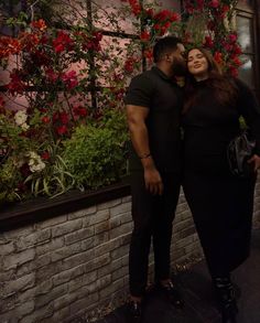 a man and woman standing next to each other in front of red flowers on a brick wall