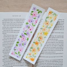 two bookmarks with watercolor flowers on them sitting on top of an open book