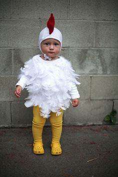 a baby dressed in a chicken costume standing next to a brick wall and wearing yellow rubber boots