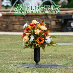 a tall black vase filled with flowers on top of a grass covered park bench next to a fountain