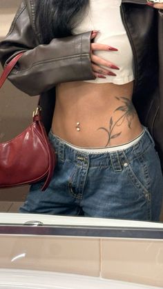 a woman with tattoos on her stomach holding a handbag and looking in the mirror