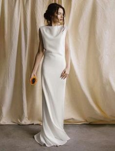 a woman standing in front of a curtain wearing a white dress and holding an orange flower