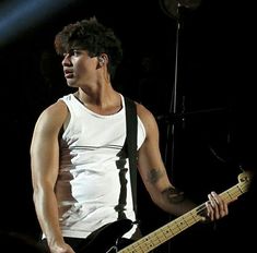 a man holding a bass guitar in his right hand and wearing a white tank top