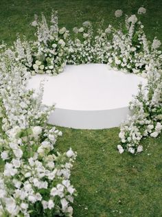 a circular white table surrounded by flowers and greenery on the grass in front of it