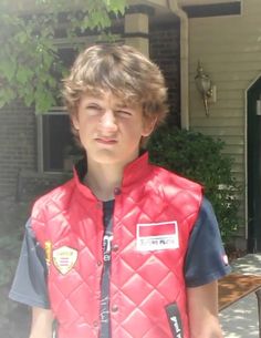 a young boy standing in front of a house wearing a red vest and blue shirt