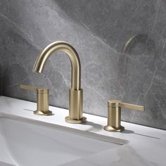 two gold faucets sitting on top of a white sink next to a marble wall