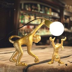 two gold monkey figurines on a wooden table with a lamp in the background