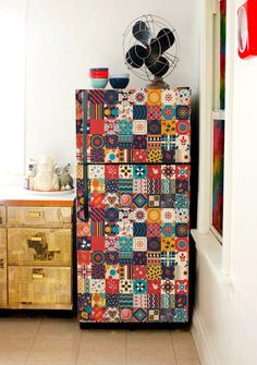 a refrigerator covered in colorful patchwork tiles next to a dresser with drawers and a fan on top