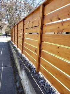 a long wooden fence next to a paved walkway with rocks on the side and trees in the background