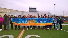 a group of people standing on top of a football field holding a large banner that says senior sunrise