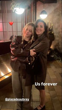 two women hugging each other in a room with lights on the ceiling and a sign that says us forever