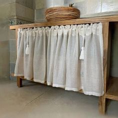 a wooden shelf with white curtains hanging on it's sides and a basket sitting on top of it