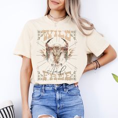 Embrace your untamed spirit with our captivating T-shirt design, a fusion of Western ruggedness and Boho charm. A majestic cow skull, intricately adorned with vibrant Boho motifs, rests upon a bed of ethereal white blooms and lush foliage, illuminated by the warmth of a Boho sun. Above, the word 'Wild' arcs boldly in retro font, while 'And Free' gracefully follows below in script, encapsulating the essence of freedom and wanderlust. Elevate your style with this symbol of untamed beauty, perfect Fall Festival Graphic Print T-shirt, Hippie Graphic Print T-shirt For Fall, Hippie Crew Neck T-shirt For Fall, Free-spirited Cotton T-shirt With Graphic Print, Bohemian Fall Crew Neck T-shirt, Bohemian Crew Neck T-shirt For Fall, Bohemian Short Sleeve T-shirt For Fall, Fall Bohemian Crew Neck T-shirt, Free-spirited Cotton Crew Neck T-shirt