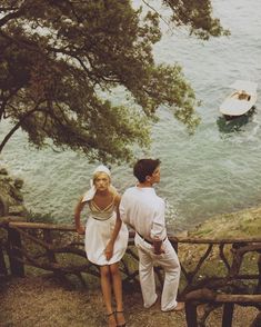 a man and woman standing next to each other on a hill overlooking the ocean with boats in the water