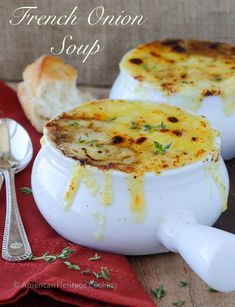 two white bowls filled with french onion soup on top of a red cloth next to silver spoons