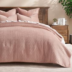 a bed with pink bedspread and pillows in a neutral room next to a potted plant