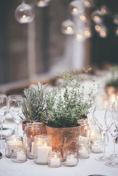 a table topped with lots of candles and vases filled with greenery next to wine glasses