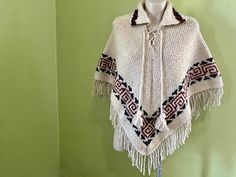 You will love how cosy and cool this vintage handmade poncho is! A chunky knit wool poncho in cream with decorative brown stripes. It has fabulous boho vibes with its turn-down collar, fringed hem and lace-up neck with pom-poms. This is a unisex style that will look amazing on anyone. Wear with a pair of denim bell bottoms for the ultimate retro style.  One size fits most. Measurements:                                                                                                                                              Widest point laying flat - 82 cm / 32 inches                                                                                           Length including fringe - 80 cm / 31 inches This item is one of a kind. BUYING VINTAGE: While all care is taken to state any obvious f Hand Knitted White Poncho For Winter, White Handmade Bohemian Poncho, Handmade Brown Bohemian Poncho, Hand Knitted Beige Poncho For Fall, Handmade Beige Poncho One Size, Traditional White Poncho For Fall, Bohemian One Size Cream Poncho, White Bohemian Poncho For Winter, Cream Bohemian Poncho For Fall