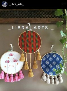 three hand embroidered hoops with tassels hanging on a wall next to a potted plant