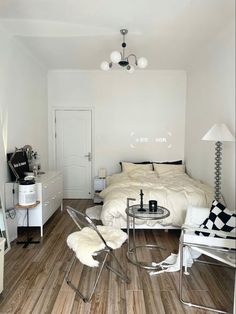 a bedroom with white walls and wood flooring has a bed, chair, nightstands, table, mirror and lamp on the wall