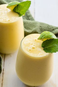 two glasses filled with yellow smoothie on top of a white wooden table next to green napkins