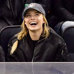 a smiling blonde woman wearing a black jacket and hat at a baseball game with other people in the stands