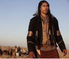 a man with long hair and braids standing in the middle of a field next to horses