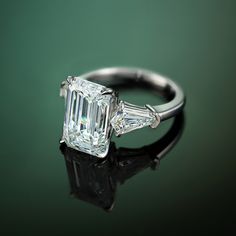 an emerald and diamond engagement ring on a reflective surface with reflection in the foreground