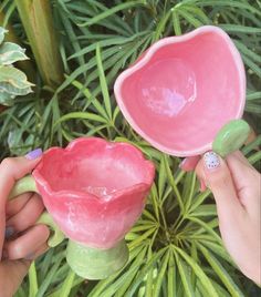 two hands are holding pink ceramic bowls in front of green leaves and plants, one has a small white dot on the end of the bowl