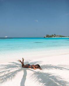 a woman laying on top of a white sandy beach next to the ocean with a boat in the background