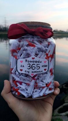 someone holding up a jar filled with red and white confetti in front of a body of water