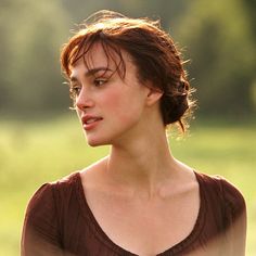 a woman standing in a field with her head turned to the side, wearing a brown top